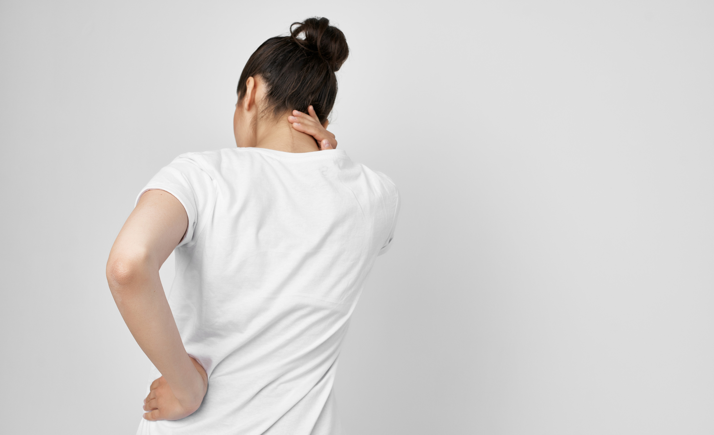 A woman in a white t-shirt seen from behind, holding her neck and lower back, indicating discomfort and body aches.