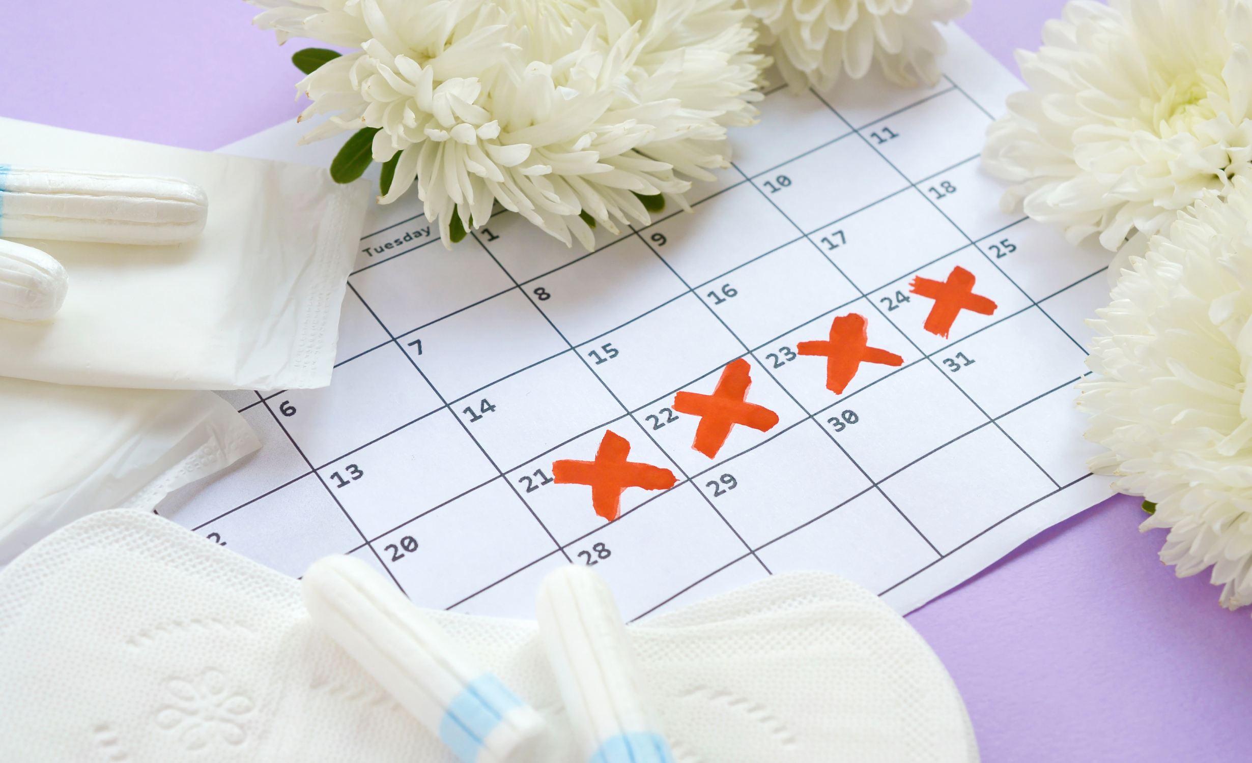 A calendar with red X marks on specific dates, surrounded by menstrual pads, tampons, and white flowers, representing irregular menstrual cycle conditions.