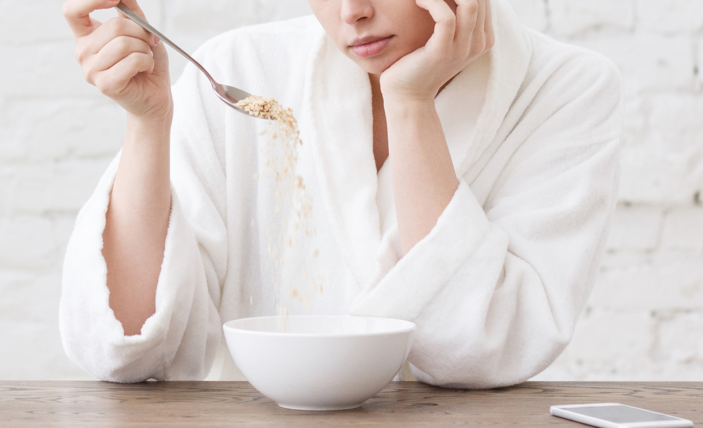 A person in a white robe sitting at a table, looking disinterested as they slowly pour cereal into a bowl, illustrating symptoms of loss of appetite conditions.