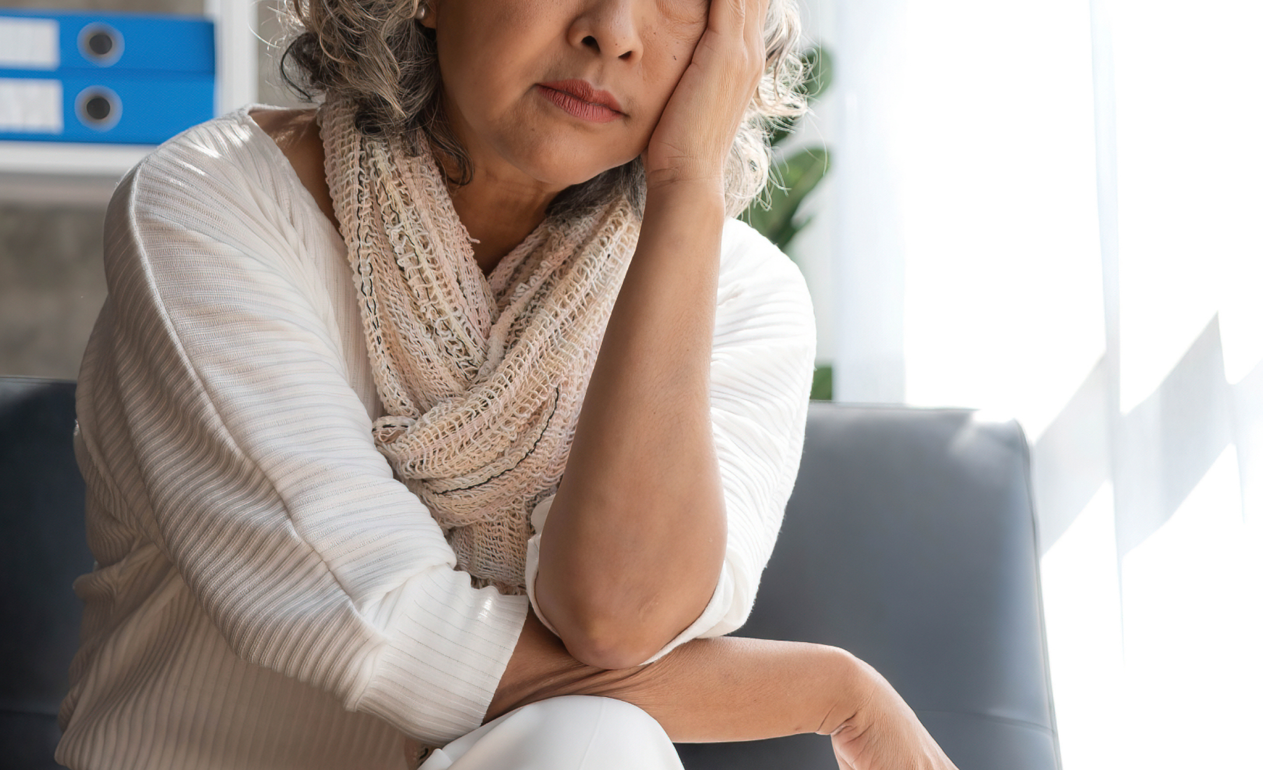 A mature woman sitting on a couch, holding her head with a concerned expression, depicting symptoms of menopause.