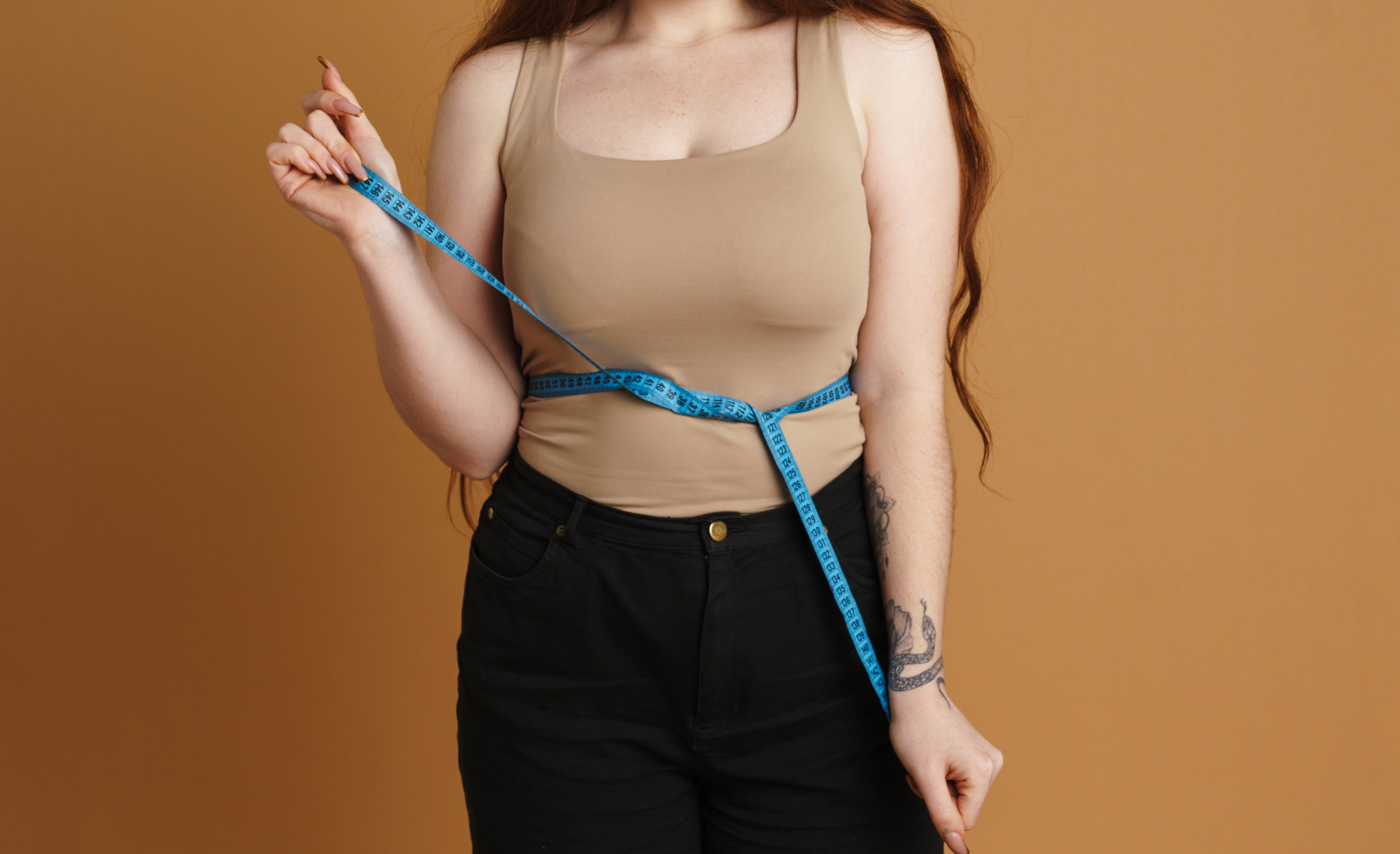 A person in a beige tank top and black pants holding a measuring tape around their waist, depicting symptoms of slowed metabolism conditions.